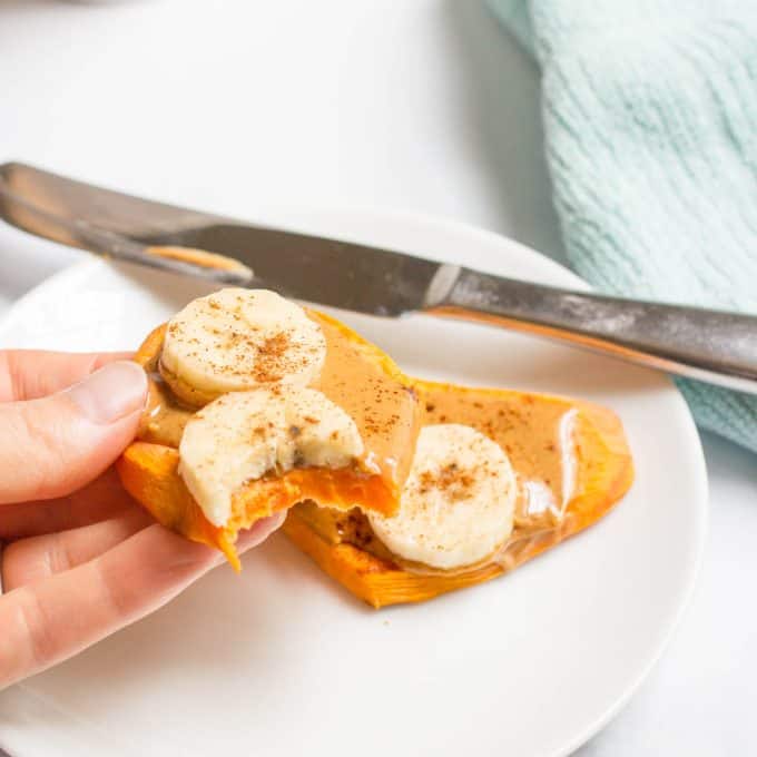 A hand holding a half of a sweet potato toast topped with peanut butter, banana slices and cinnamon with a bite taken out.