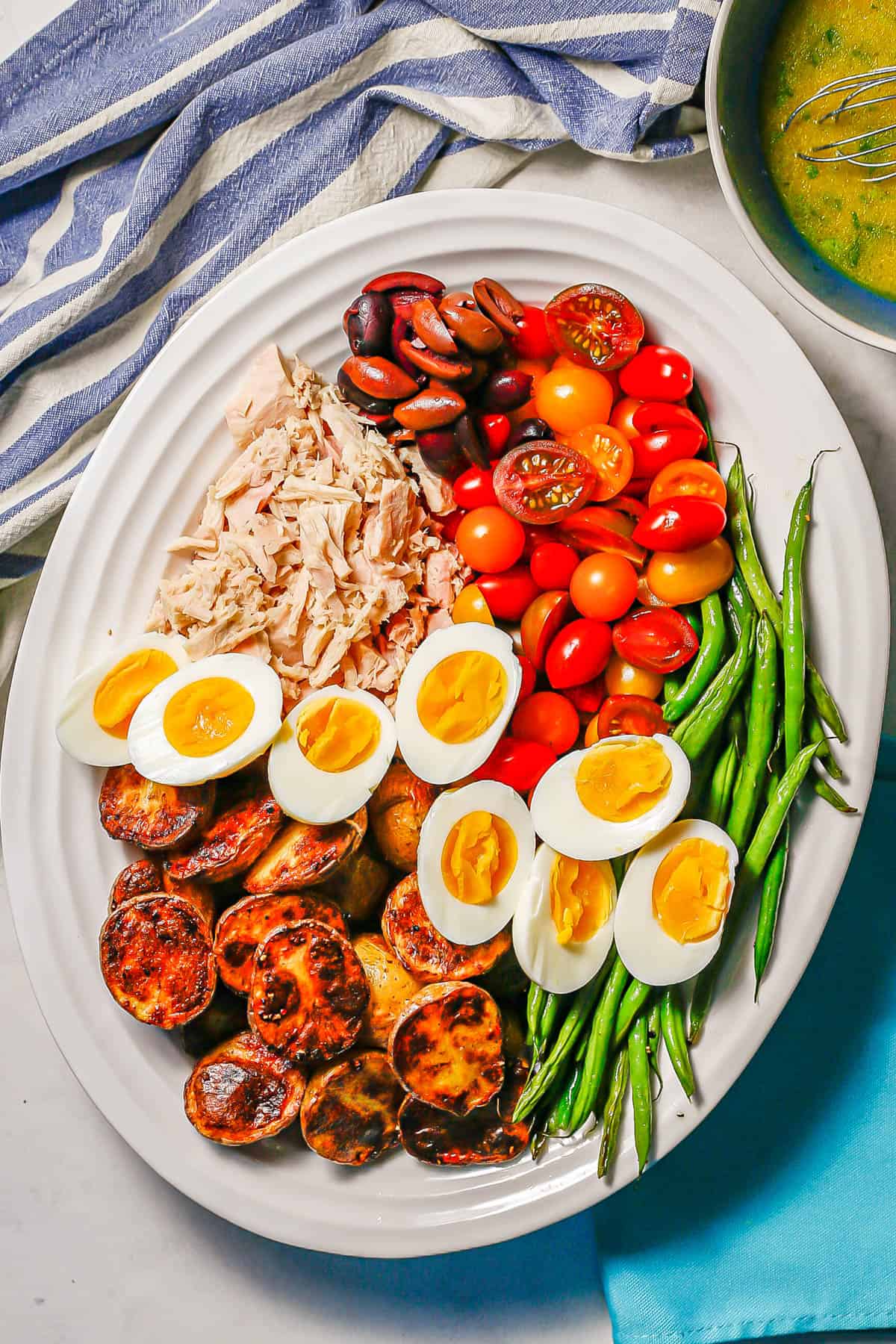 Tuna Niçoise salad with roasted potatoes and green beans, hard boiled eggs, colorful tomatoes and olives on a white platter with a lemony vinaigrette to the side.