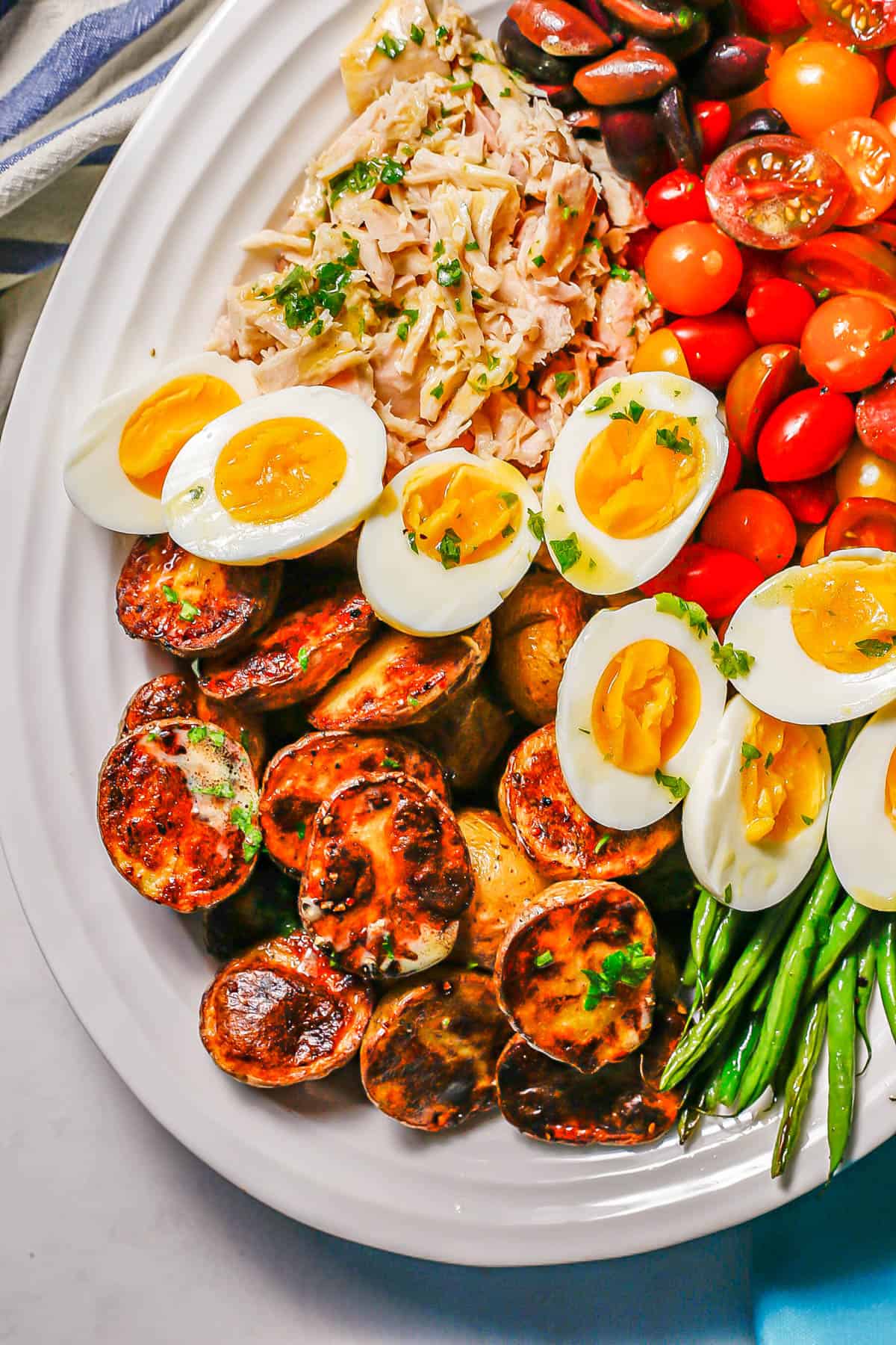 Close up of roasted potatoes, hard boiled eggs and tuna as part of a Niçoise salad served on a white platter.
