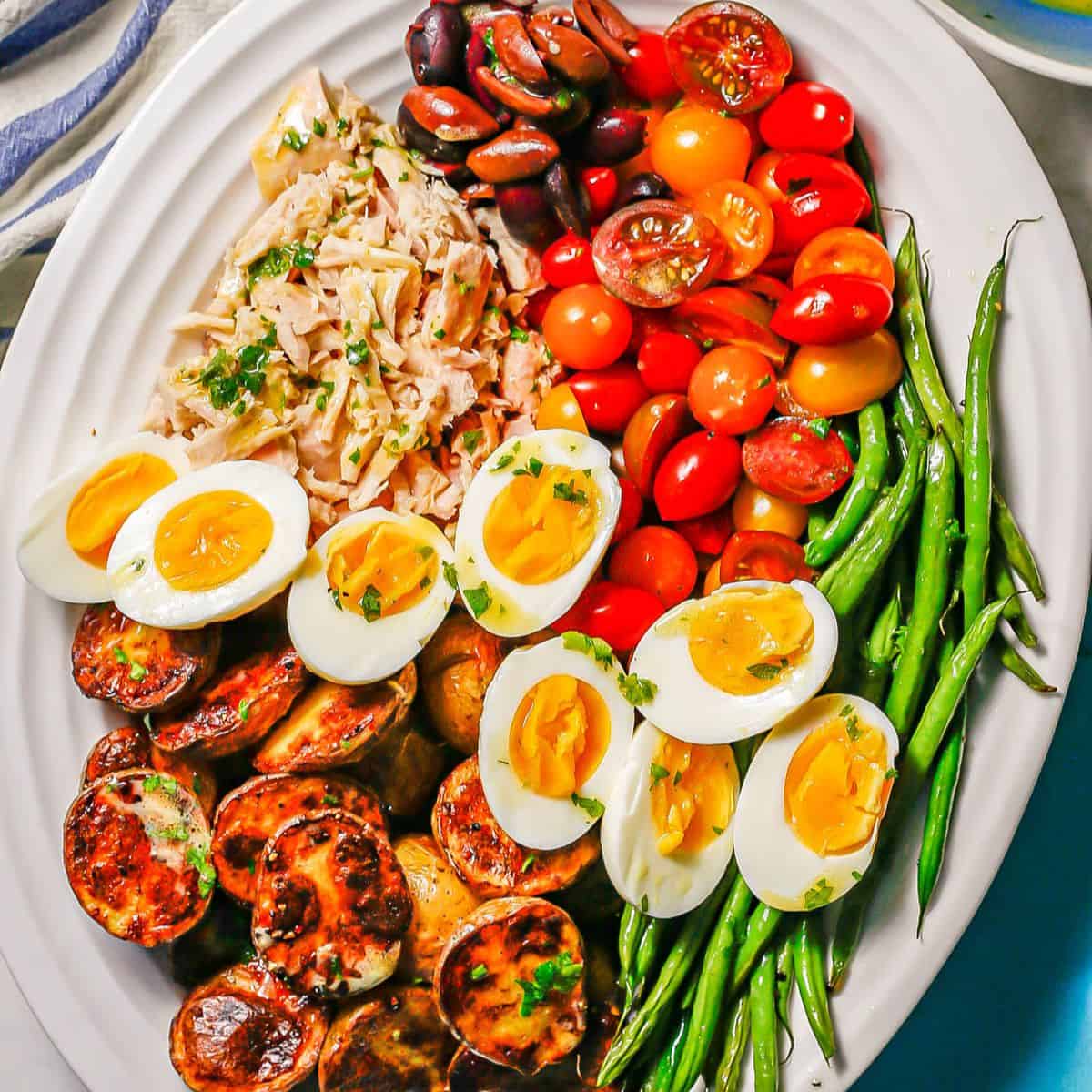 Close up of a Niçoise salad with potatoes, green beans, eggs, tomatoes, tuna and olives on a large white platter.