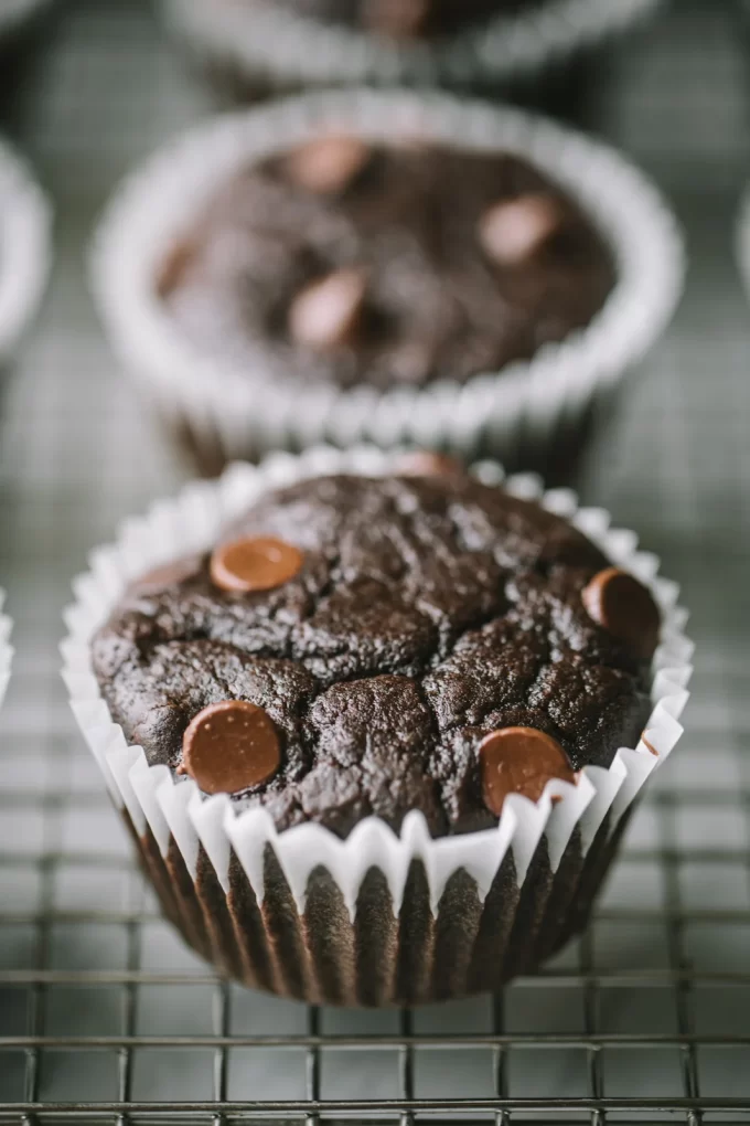 Close up of a chocolate black bean muffin with chocolate chips in a muffin wrapper on a cooling rack.