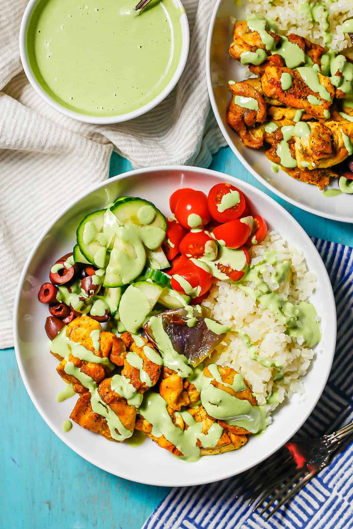 Two low white bowls with caulifower rice, turmeric chicken, fresh veggies and a tahini herb dressing drizzled over top.