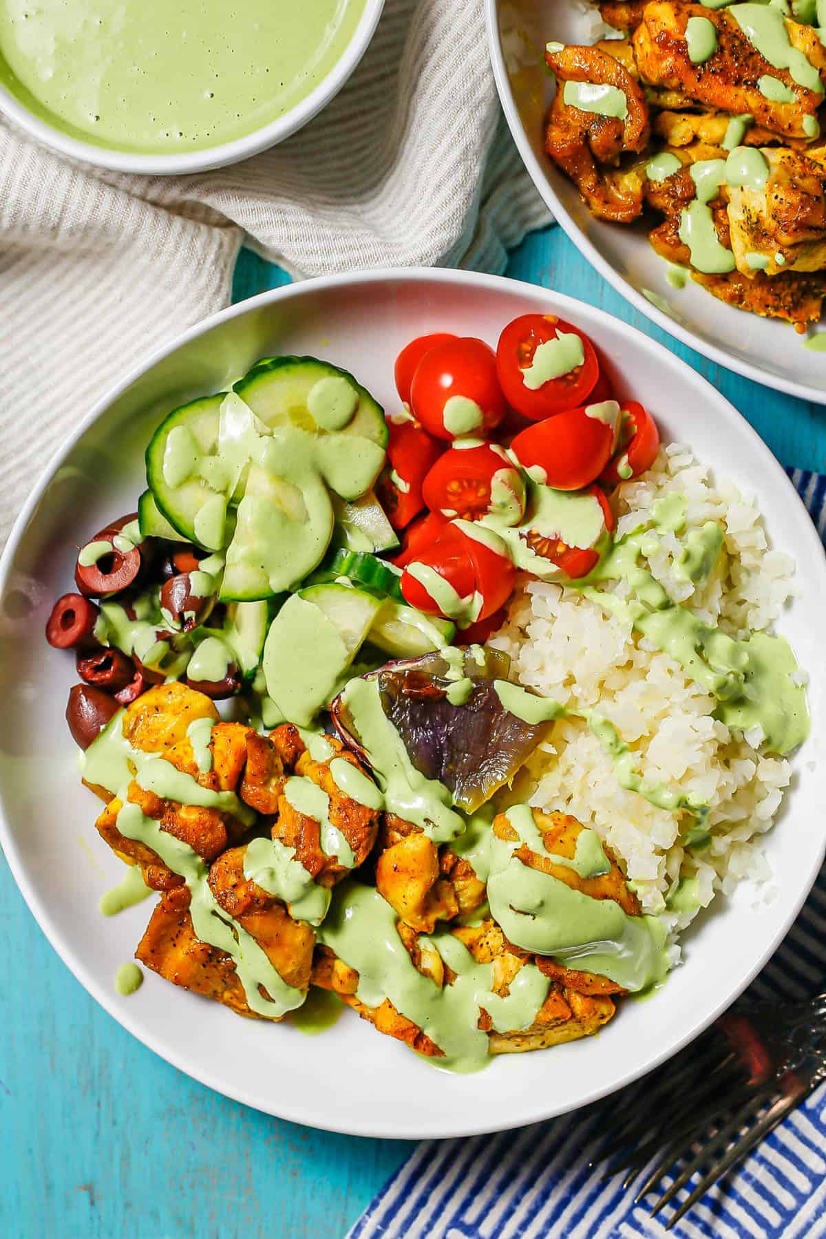 A low white bowl with caulifower rice, turmeric chicken, fresh veggies and a tahini herb dressing drizzled over top.