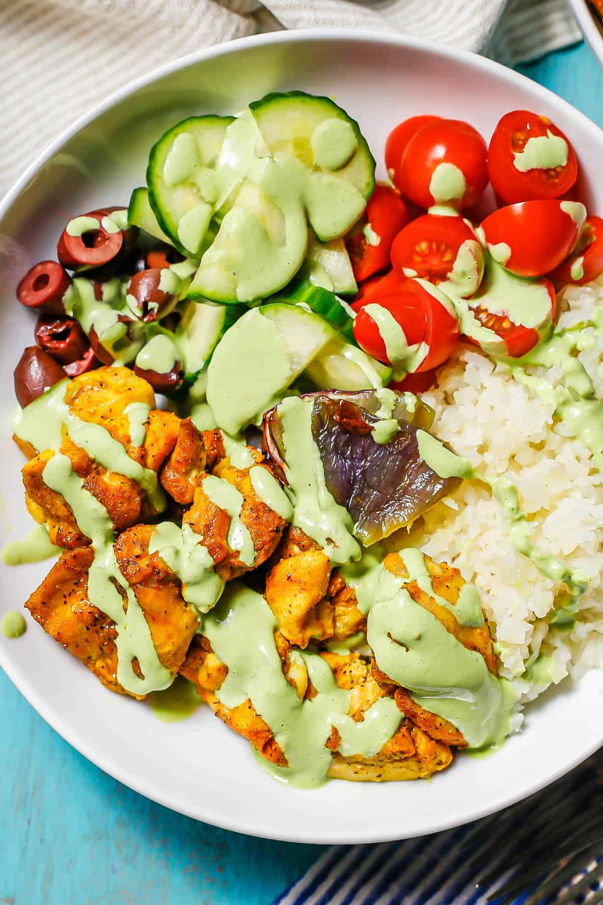 Side view of a cauliflower rice bowl with turmeric chicken thighs and fresh veggies, all drizzled with an herby dressing.