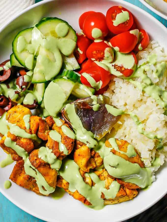 Close up of a cauliflower rice bowl with chicken, cucumbers, tomatoes and a green dressing drizzled over top.