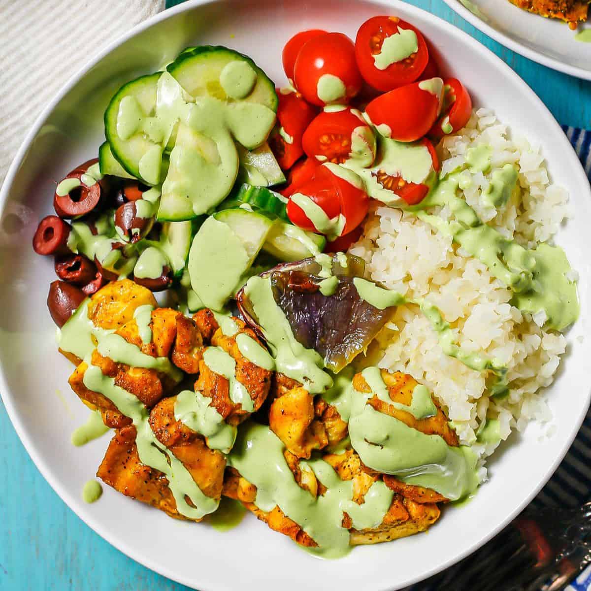 Close up of a cauliflower rice bowl with chicken, cucumbers, tomatoes and a green dressing drizzled over top.