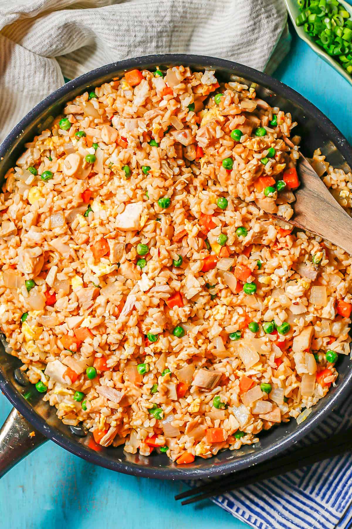 Chicken fried rice in a large dark skillet with a wooden spoon lifting a scoop and some chopped green onions in a dish to the side.