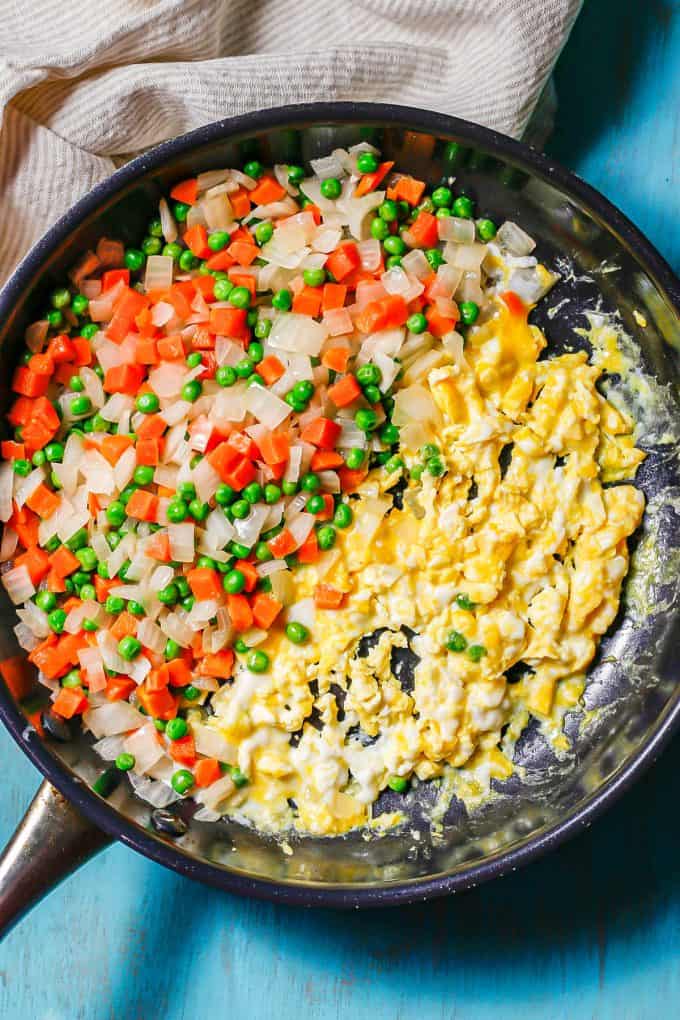 Sautéed onions with peas and carrots alongside scrambled eggs in a large dark skillet.