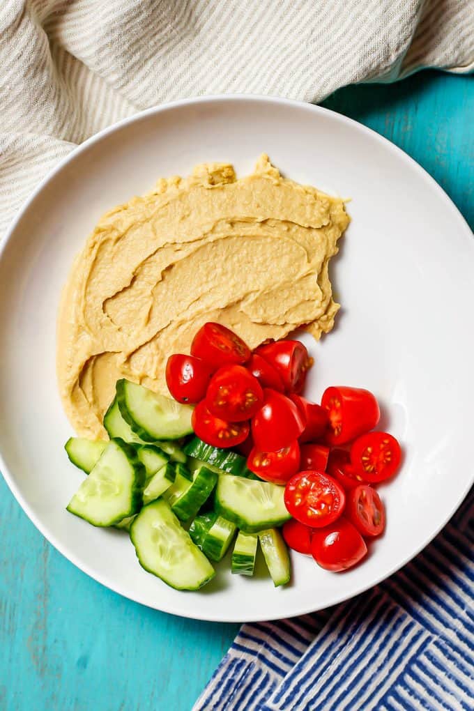 A low white bowl with a spread of hummus and cut cucumber slices and tomatoes to the side.