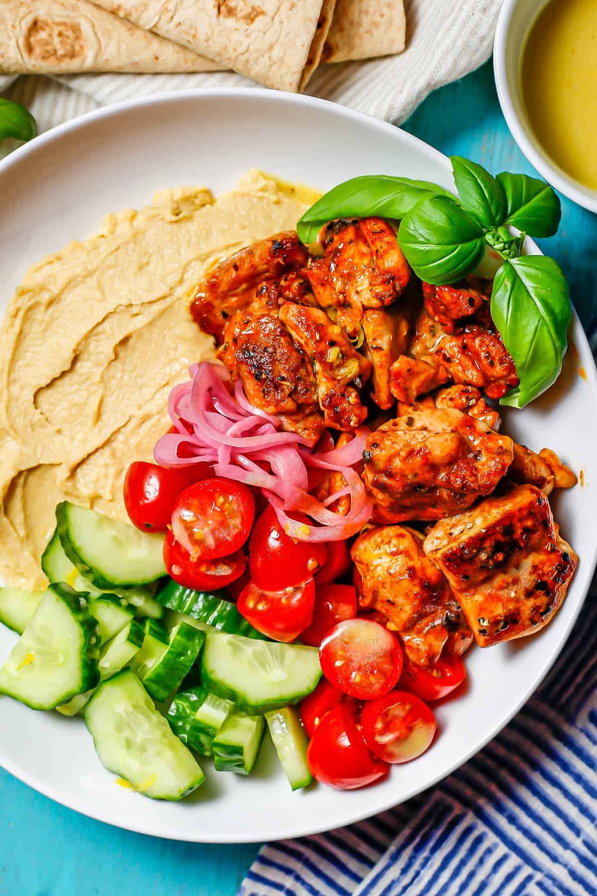 Close up of paprika chicken and hummus bowls with fresh veggies in a white bowl.