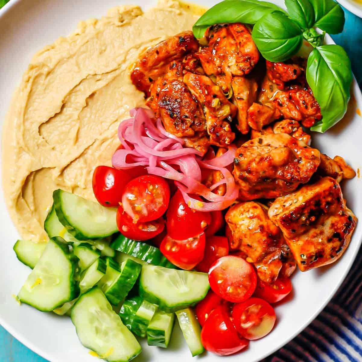 Close up of chicken hummus bowls with veggies and a basil garnish in a round white low bowl.