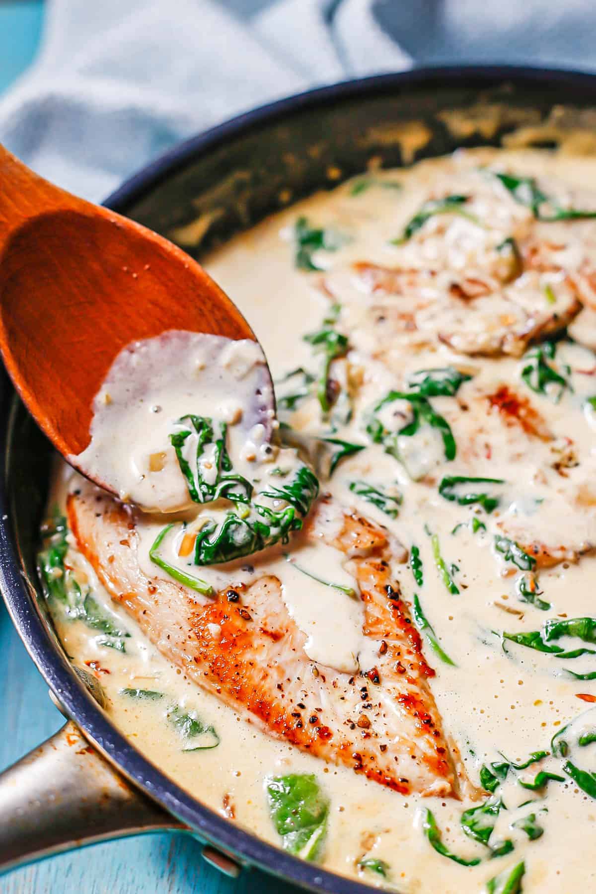 A wooden spoon adding spinach and sauce to a chicken breast in a dark skillet.