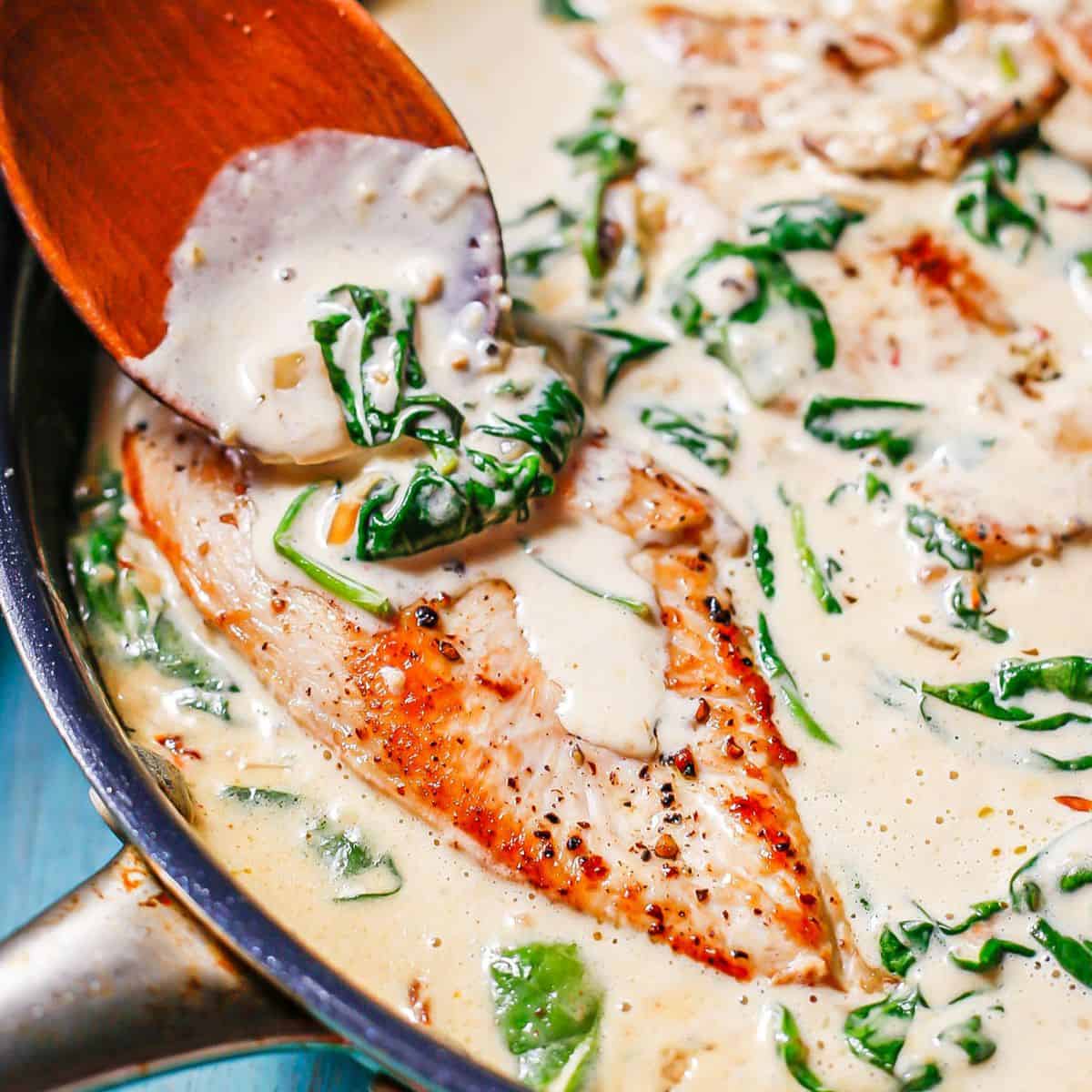 A square image of a wooden spoon adding spinach and sauce to a chicken breast in a dark skillet.