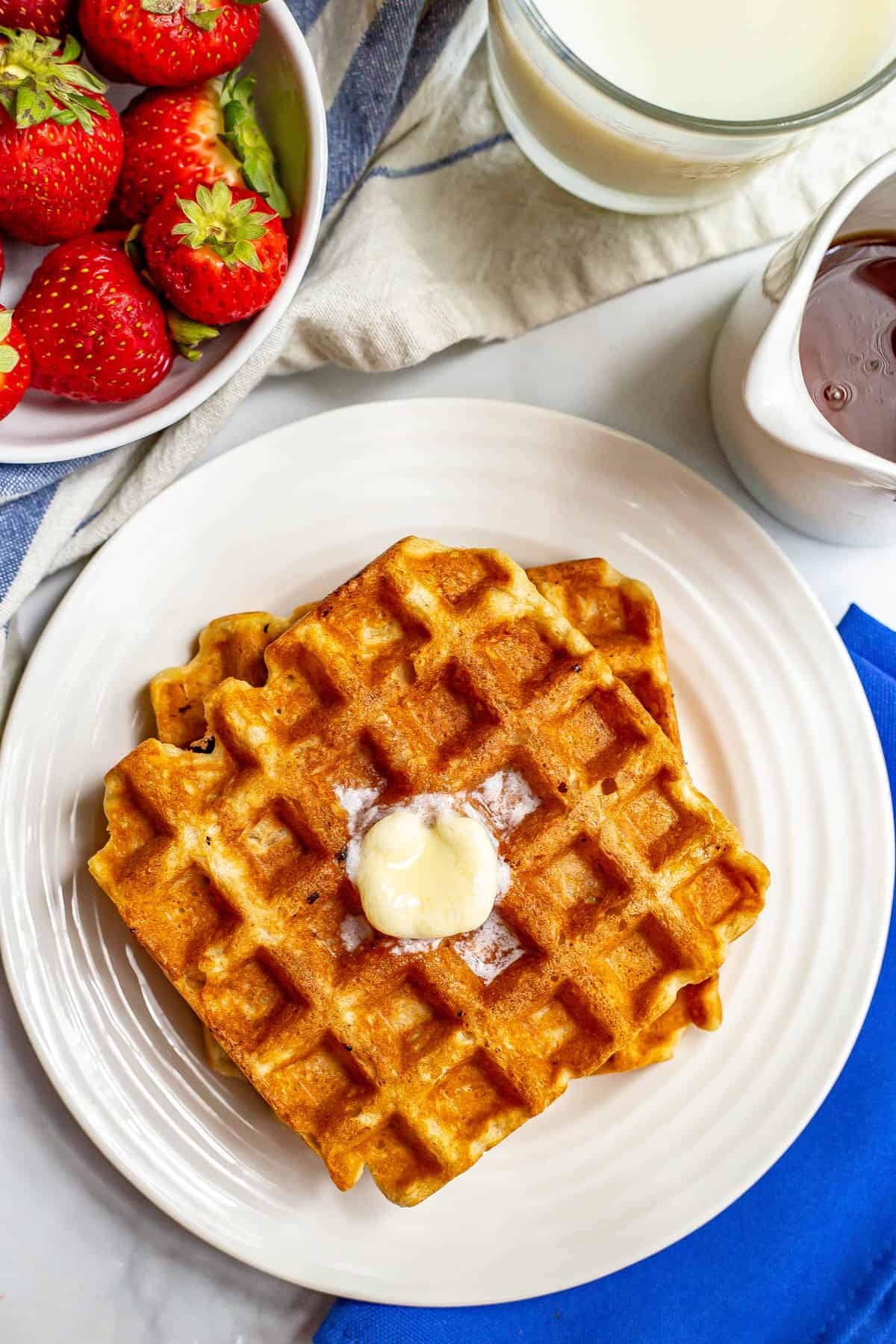 Homemade waffles stacked on a white plate with butter melting on top and syrup to the side.