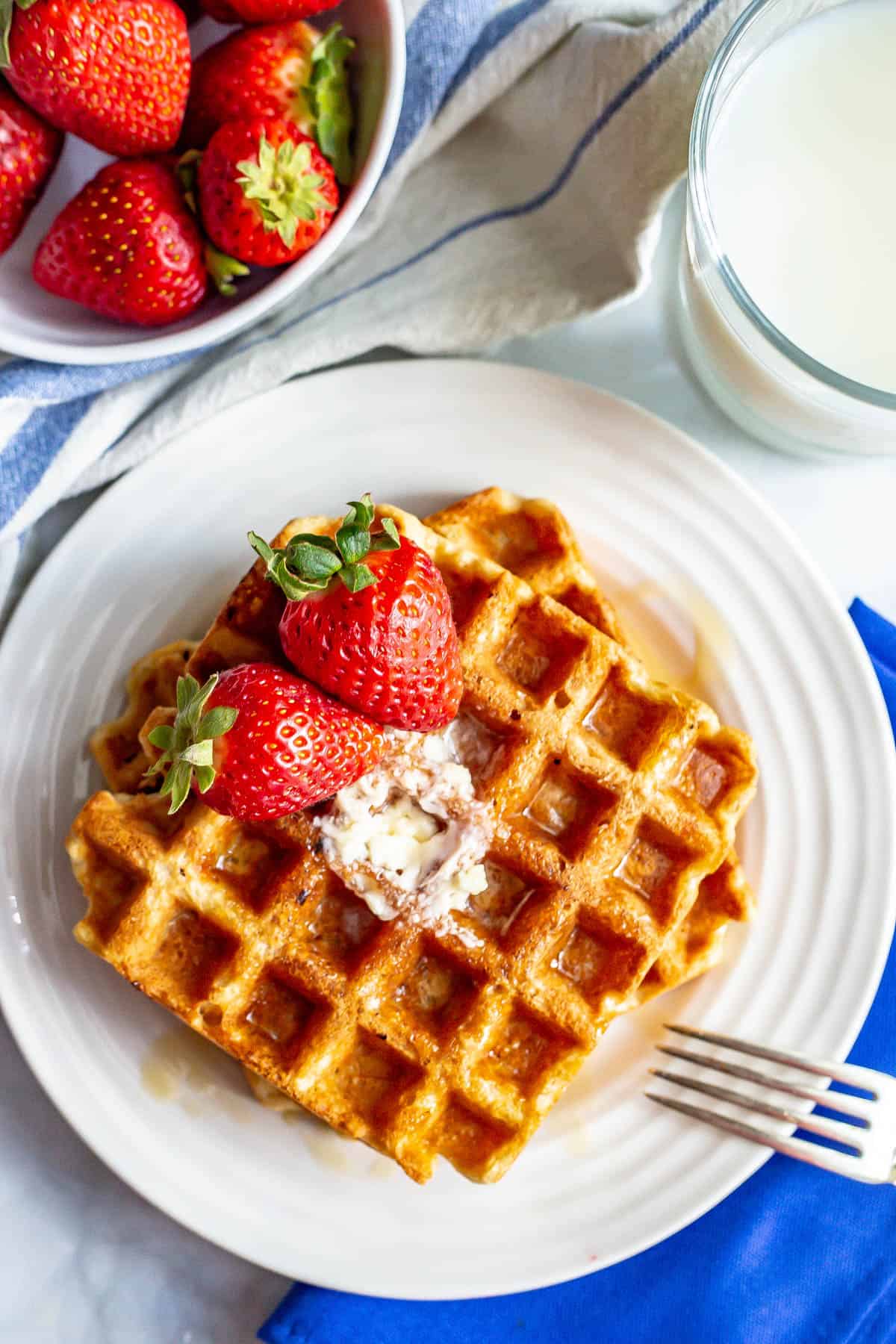 A fork resting on a plate with two whole wheat waffles topped with butter and maple syrup and two strawberries.