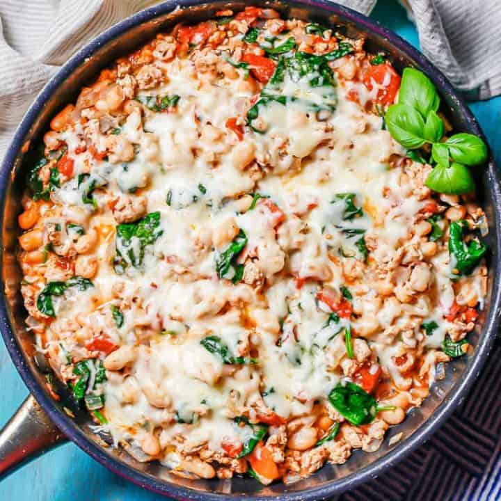 A large skillet with a cheesy ground chicken, beans and veggie mixture set on a turquoise counter with napkins to the side.
