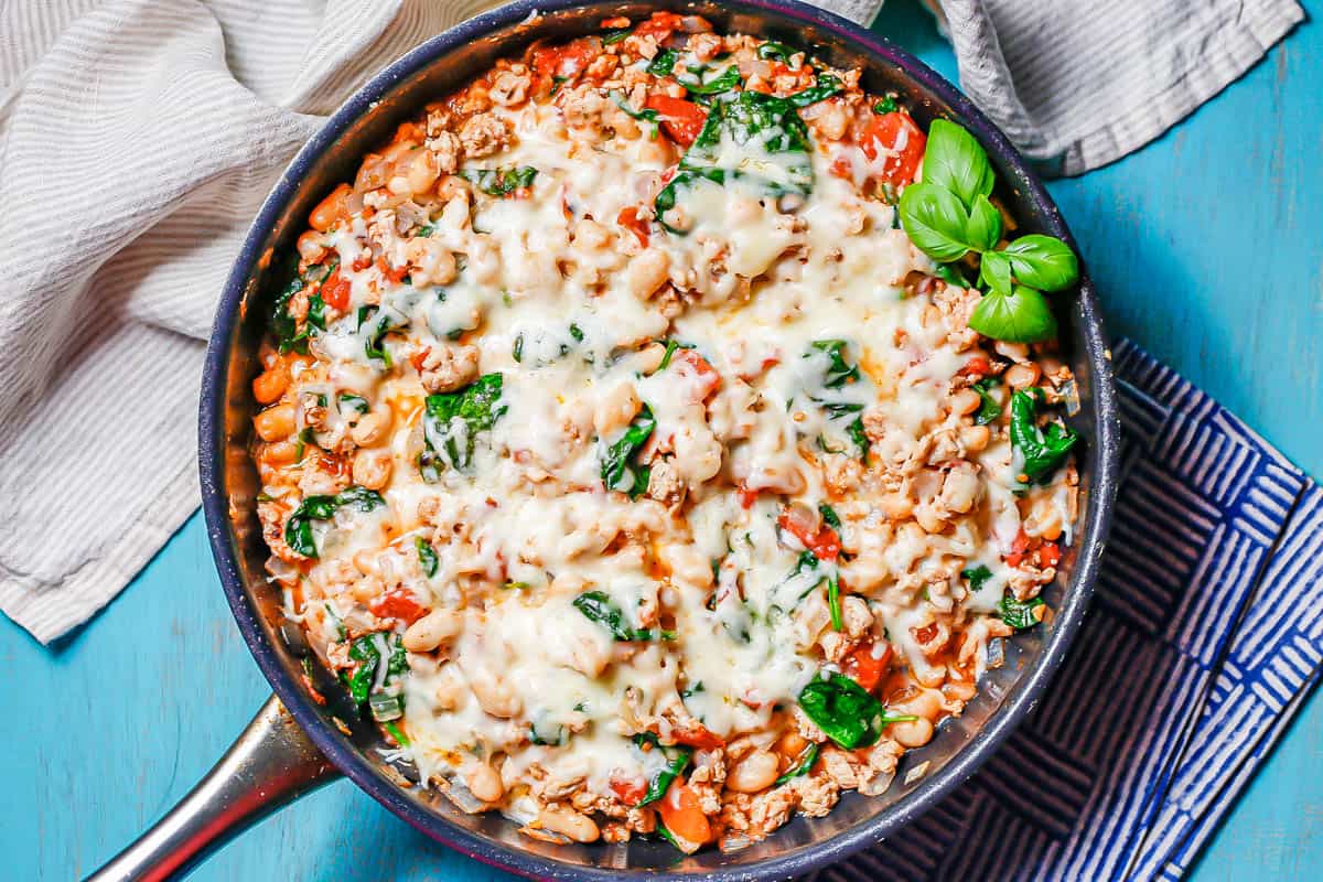 A large skillet with a cheesy ground chicken, beans and veggie mixture set on a turquoise counter with napkins to the side.