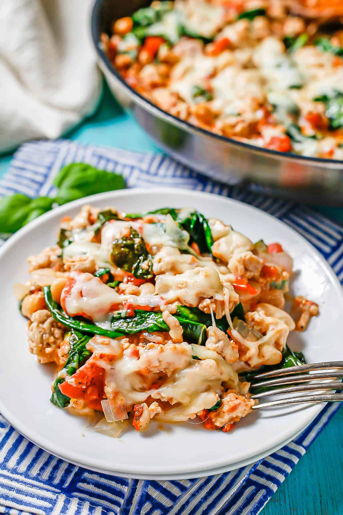 A serving of ground chicken with white beans, tomatoes and spinach, topped with cheese, on a round white plate with a fork resting on it.