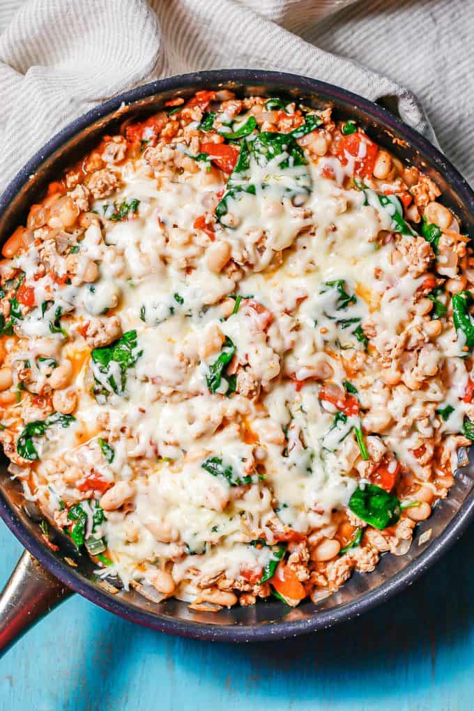 Melted mozzarella cheese on top of a mixture of ground chicken, white beans, spinach and tomatoes in a large dark pan.