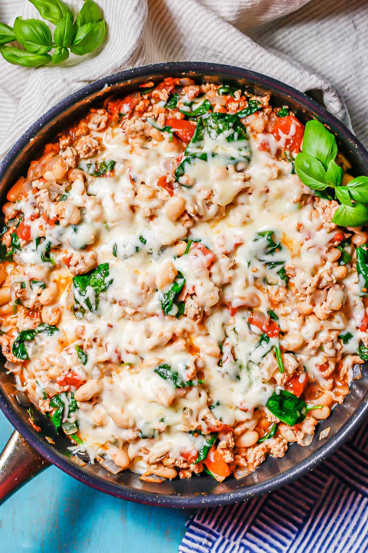 Melted mozzarella cheese on top of a mixture of ground chicken, white beans, spinach and tomatoes in a large dark pan with basil sprigs for a garnish.