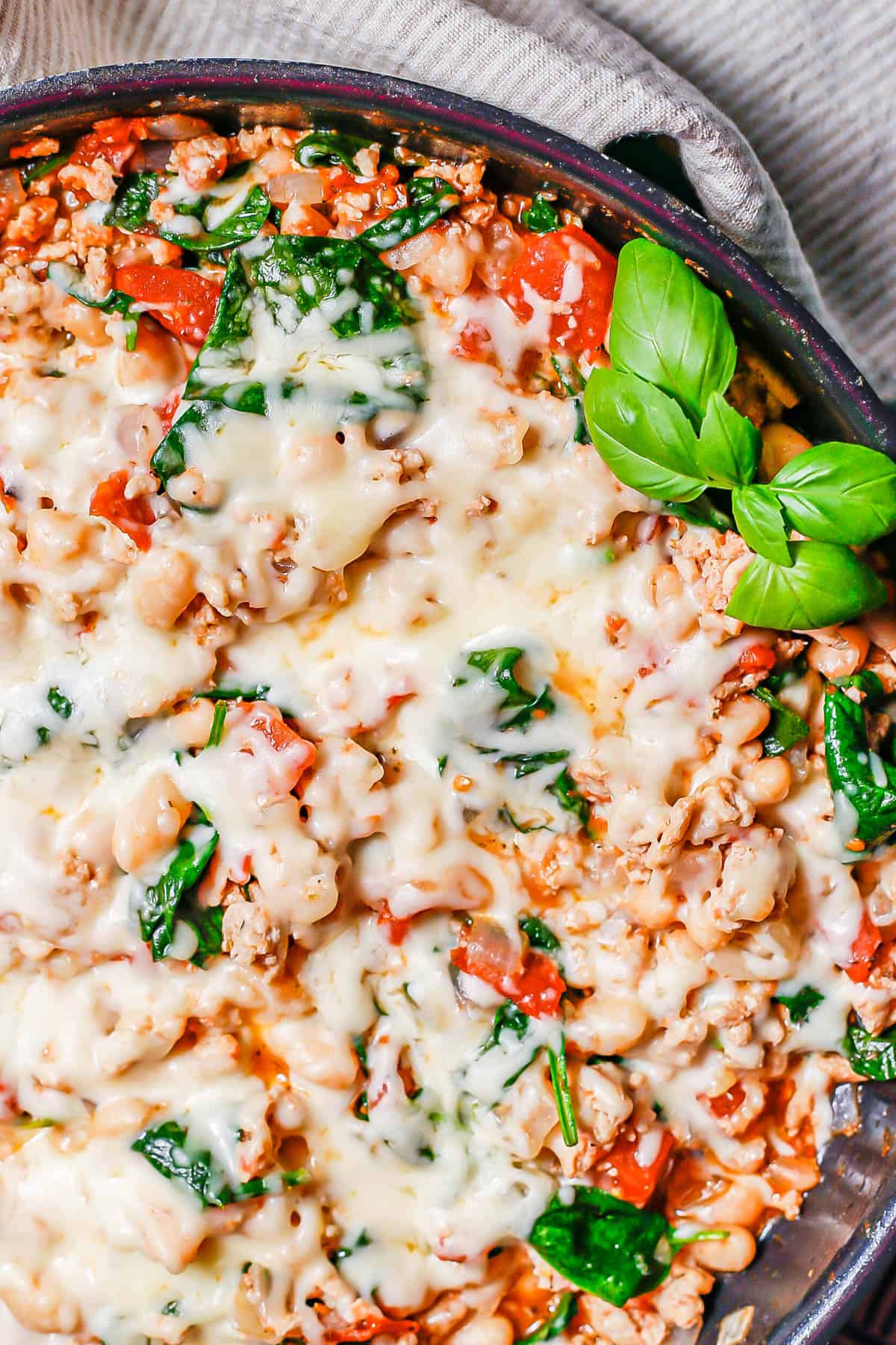 Close up of melted mozzarella cheese on top of a mixture of ground chicken, white beans, spinach and tomatoes in a large dark pan.
