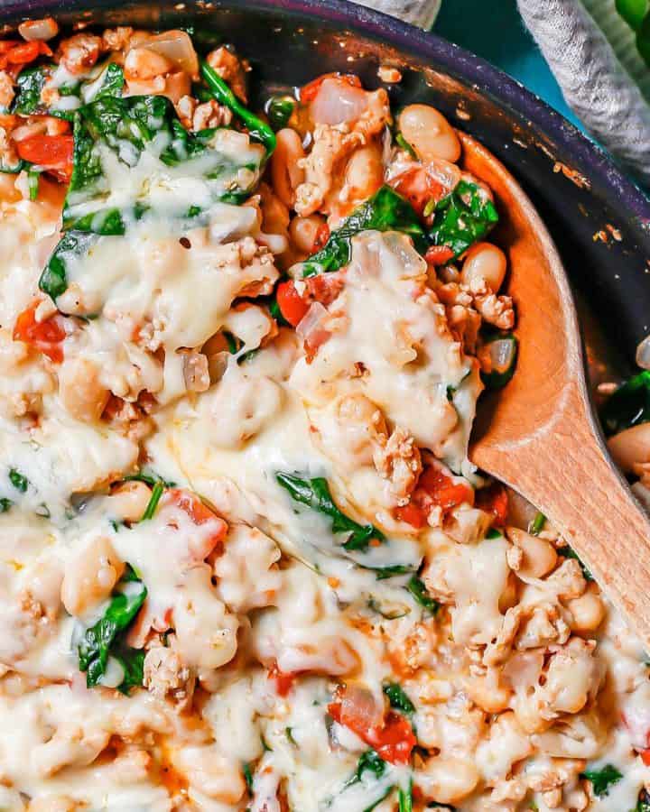 A wooden spoon scooping up a chicken, bean and veggie mixture for a large pan.