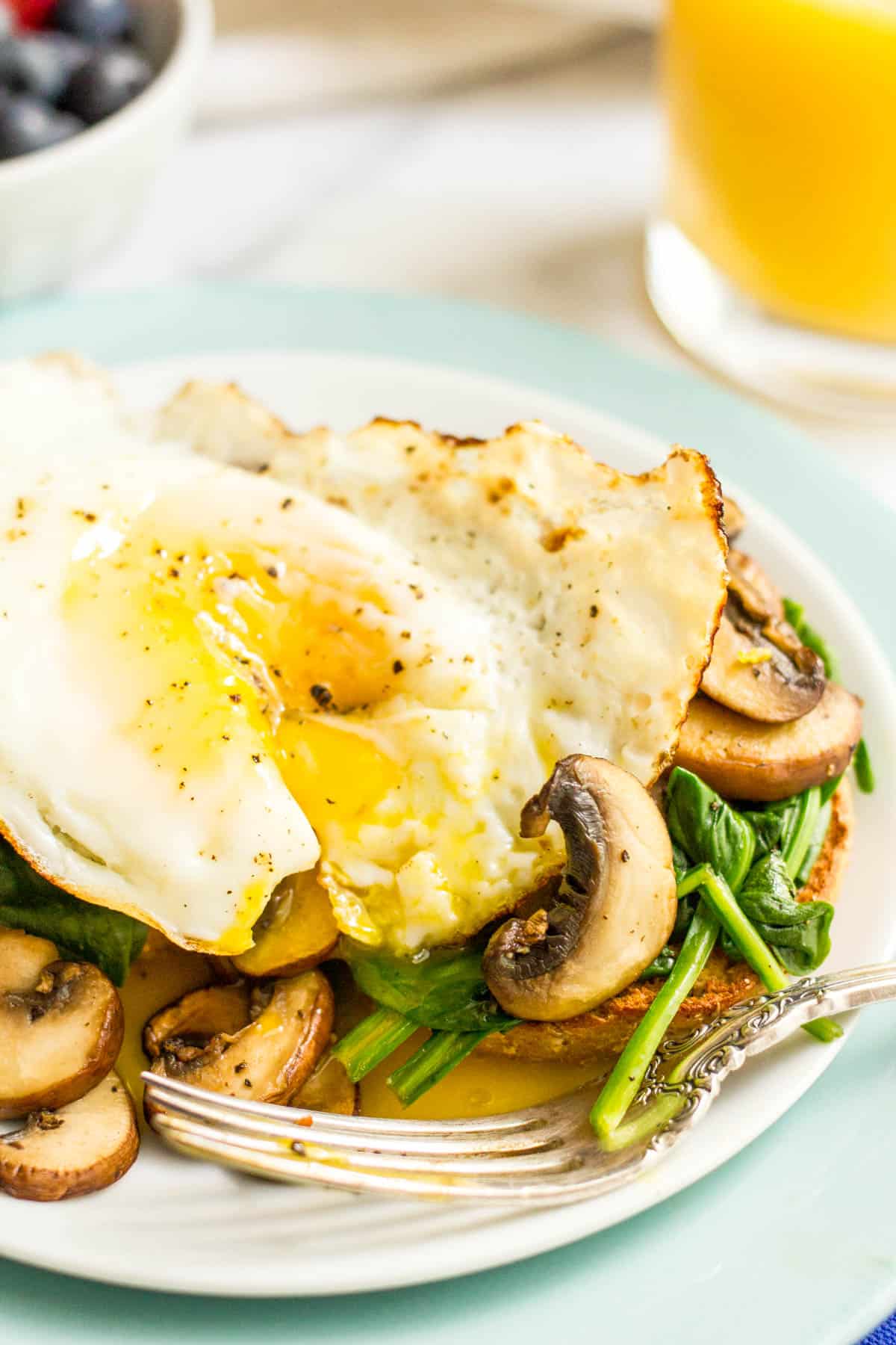 A bagel topped with sautéed spinach and mushrooms and a fried egg that has a runny yoke spilling onto the plate.