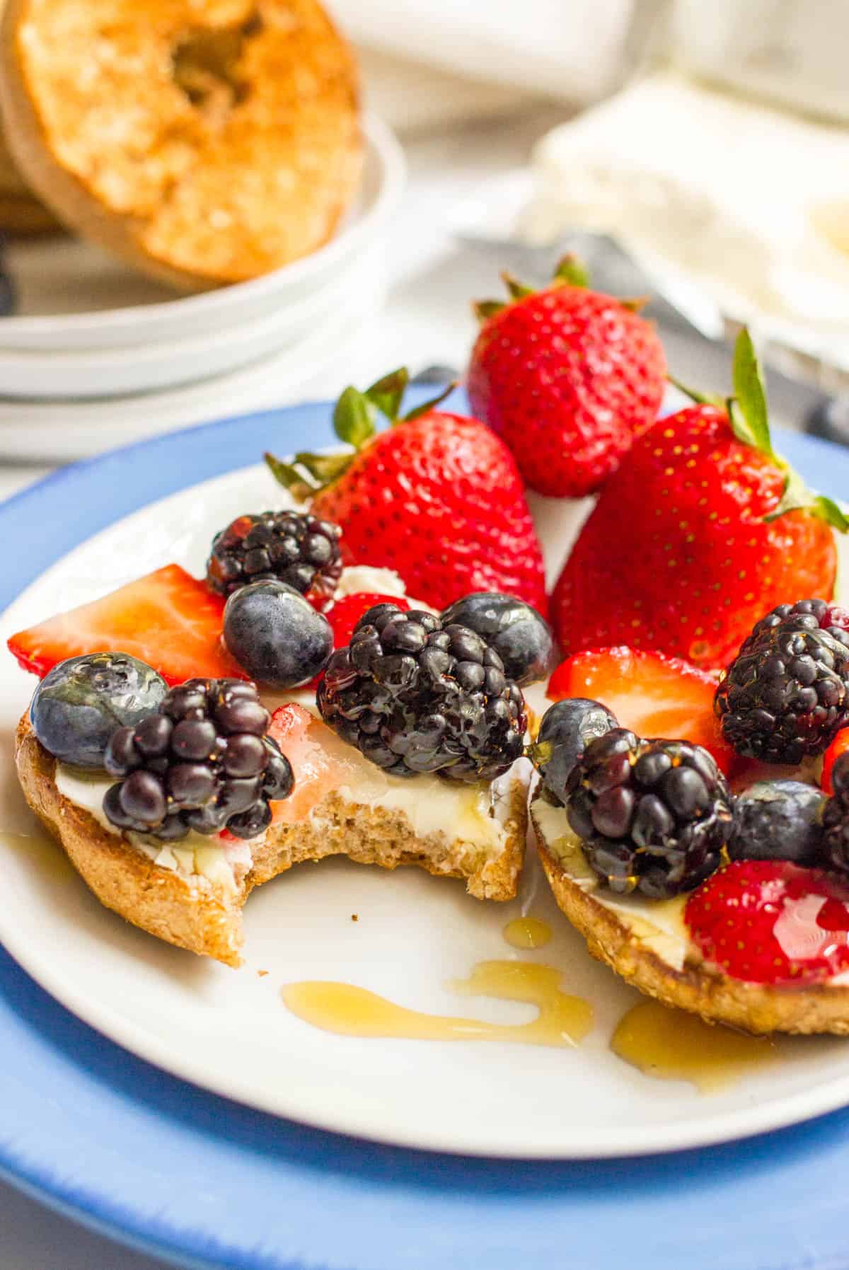 A bite taken out of a bagel topped with cream cheese, fresh berries and a drizzle of honey.