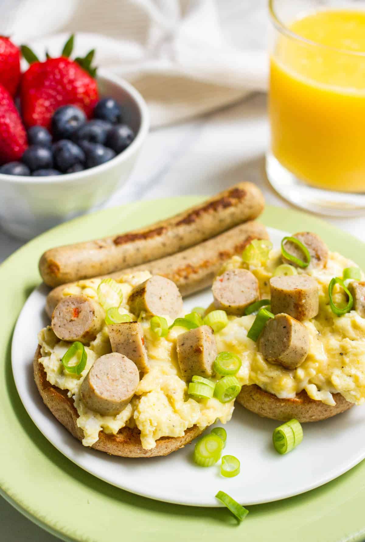 A bagel topped with scrambled eggs and sliced chicken sausages and garnished with sliced green onions.