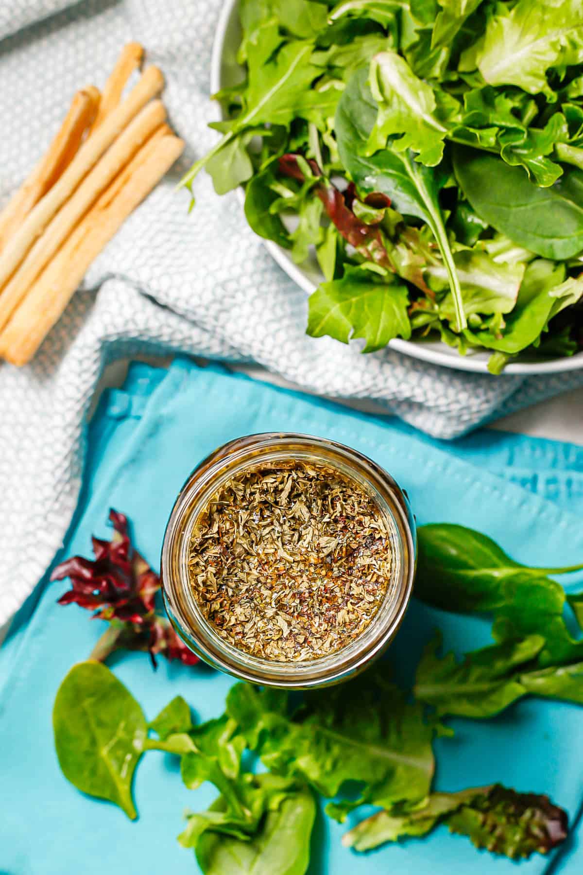 An overhead view of a glass jar of salad dressing with herbs at the top and a bowl of mixed greens to the side.