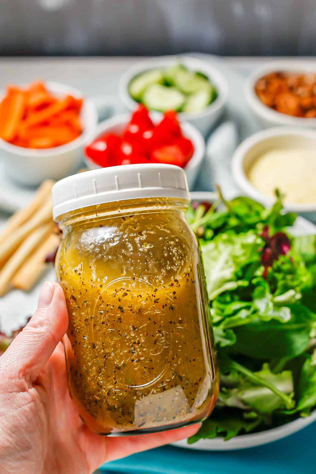A hand holding a jar of homemade salad dressing that's been shaken up with a salad spread in the background.