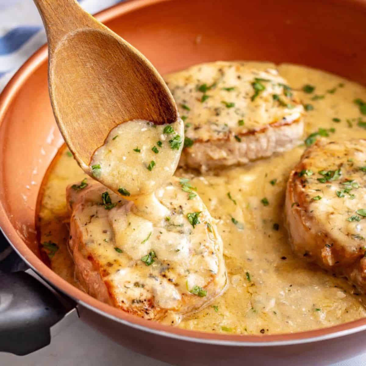 A wooden spoon drizzling gravy over a seared pork chop in a pan.