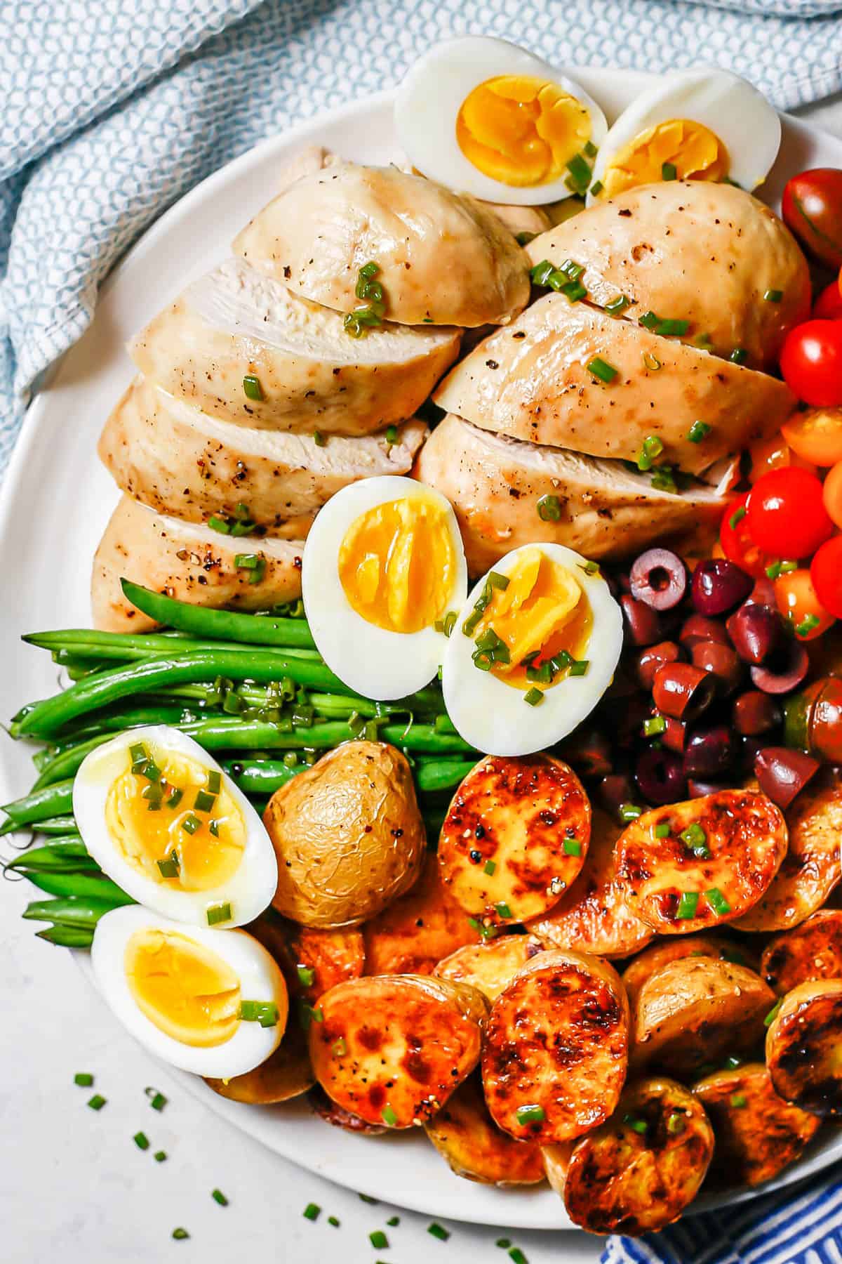 Sliced chicken breasts, hard boiled eggs, haricots verts and roasted potatoes on a white round serving plate.