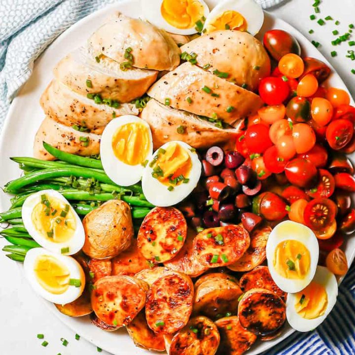 An arranged platter of roast chicken Niçoise salad with a lemony chive dressing over top on a large white plate with chives scattered nearby.