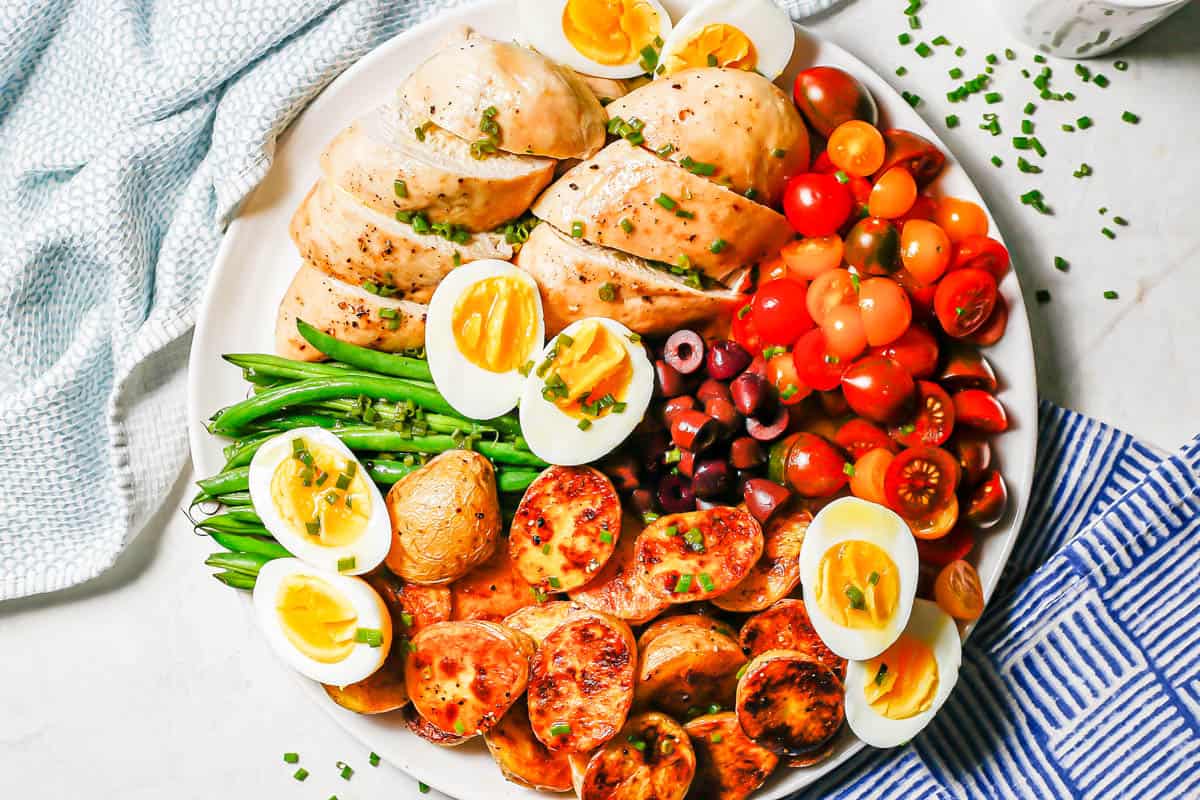 An arranged platter of roast chicken Niçoise salad with a lemony chive dressing over top on a large white plate with chives scattered nearby.