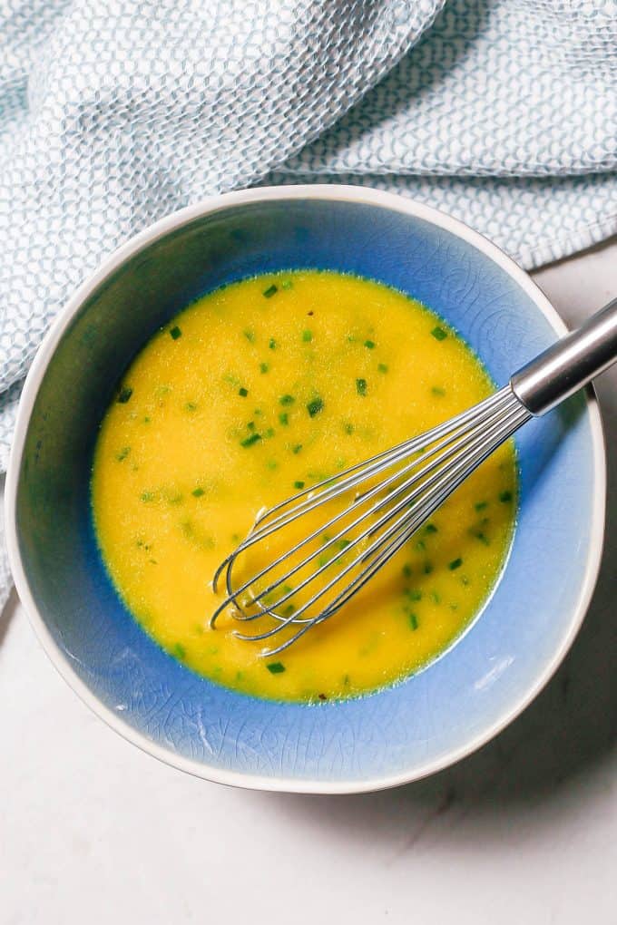 A blue and white bowl with a lemony vinaigrette dressing with chives and a whisk resting in the bowl.