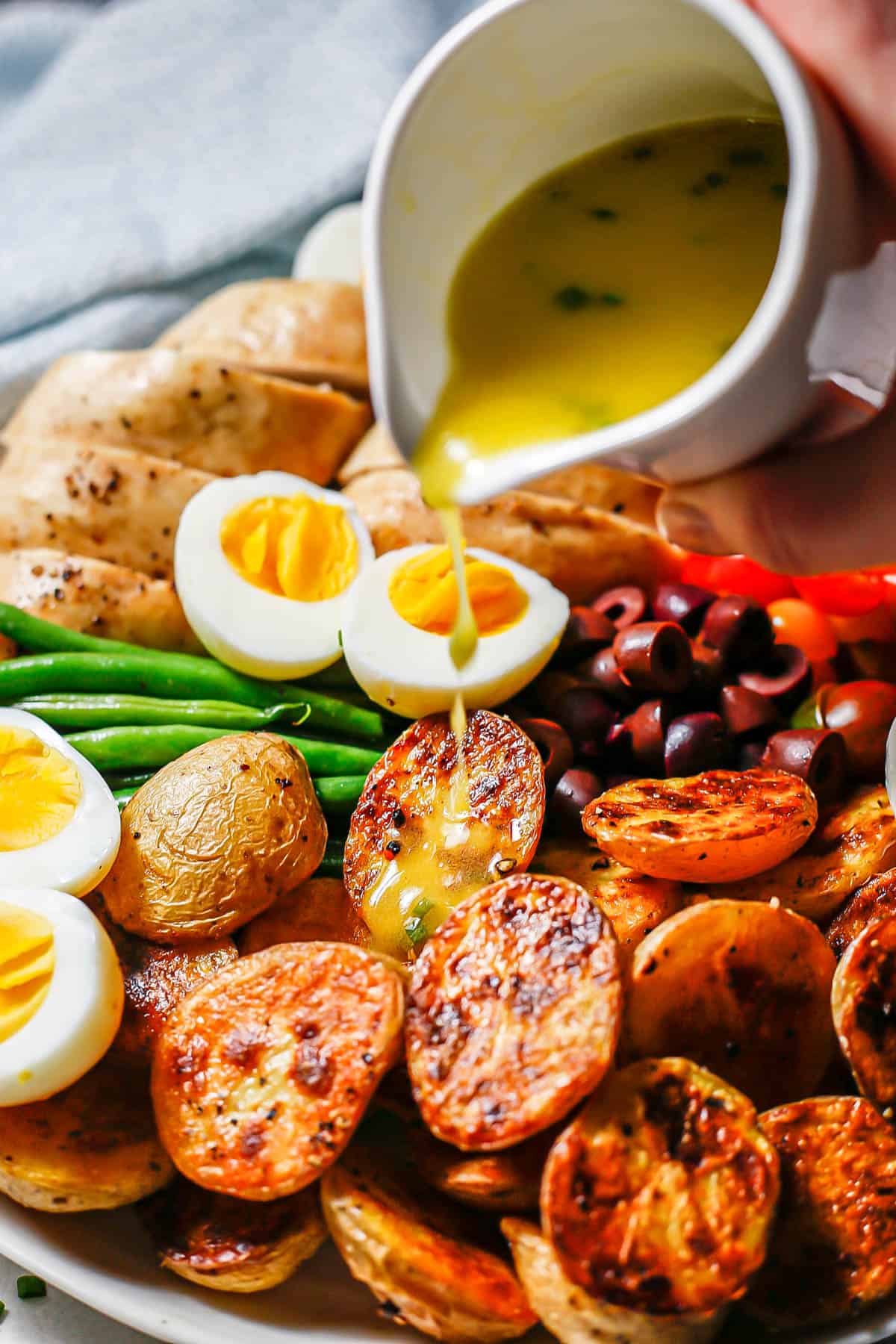 A small pitcher of dressing being drizzled over roasted potatoes on an arranged chicken Niçoise salad platter.