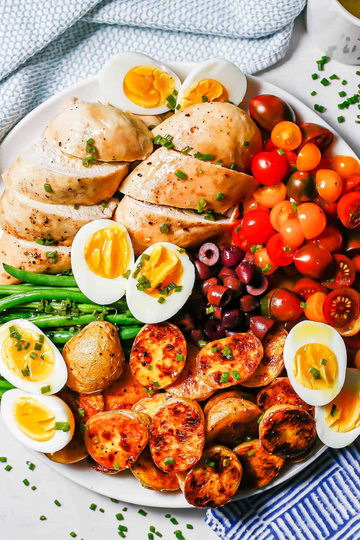 Roast chicken Niçoise salad on a large round white platter with tomatoes, olives, eggs, potatoes and haricot verts.