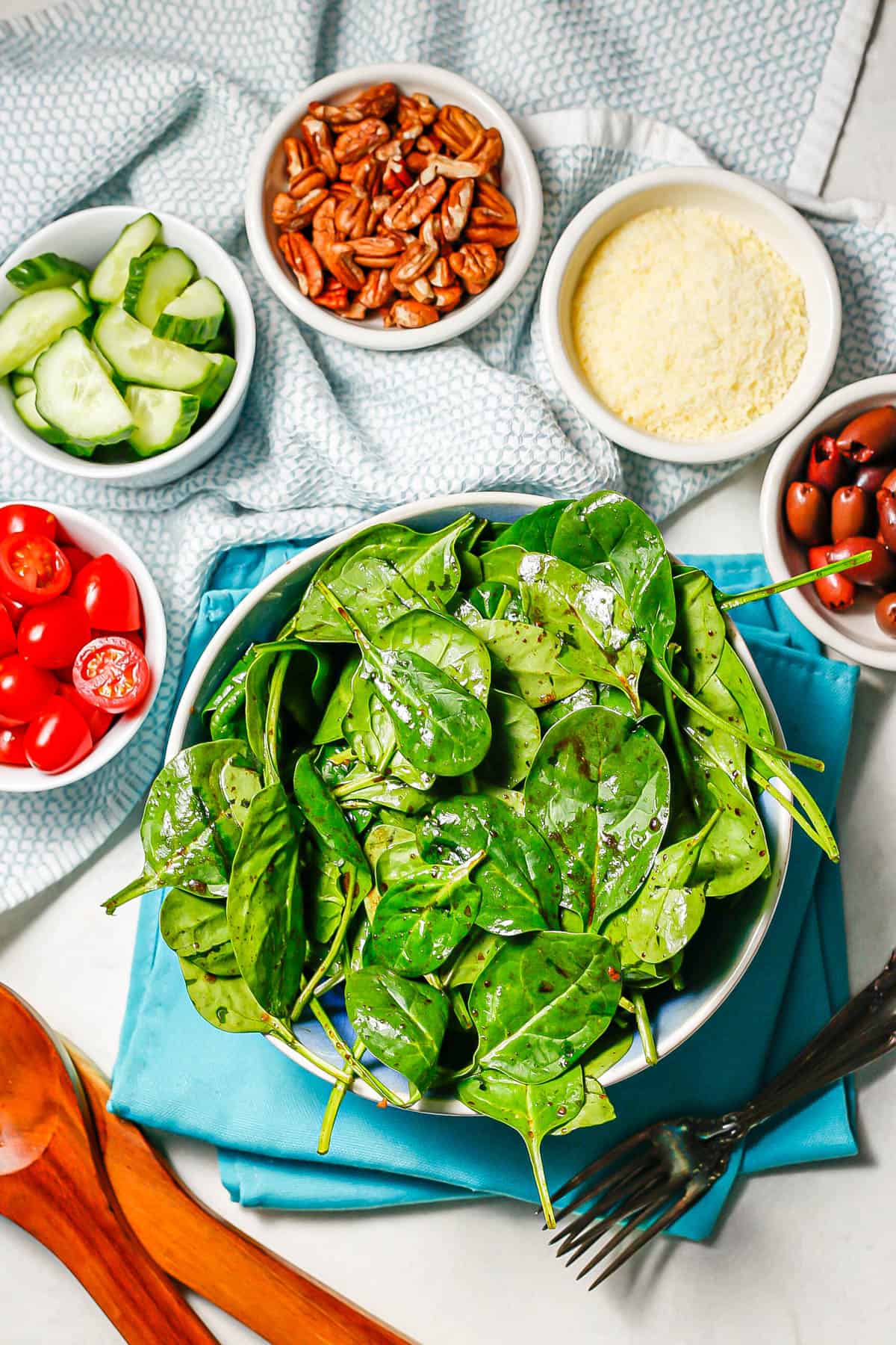 A spinach salad served in a large blue and white bowl with smaller bowls of toppings around it, including tomatoes, cucumbers, pecans, Parmesan cheese and olives.