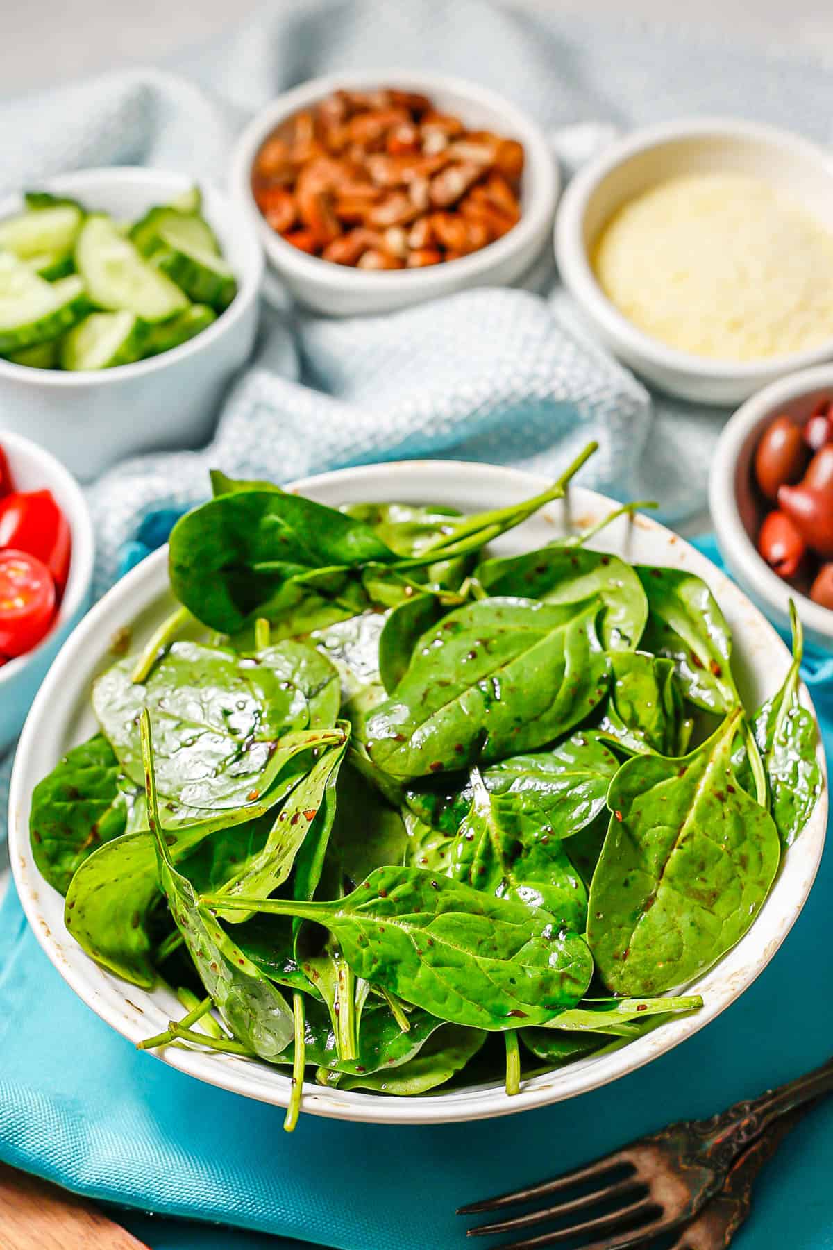 A salad bowl with spinach dressed in balsamic vinaigrette with other salad toppings in individual bowls in the background.