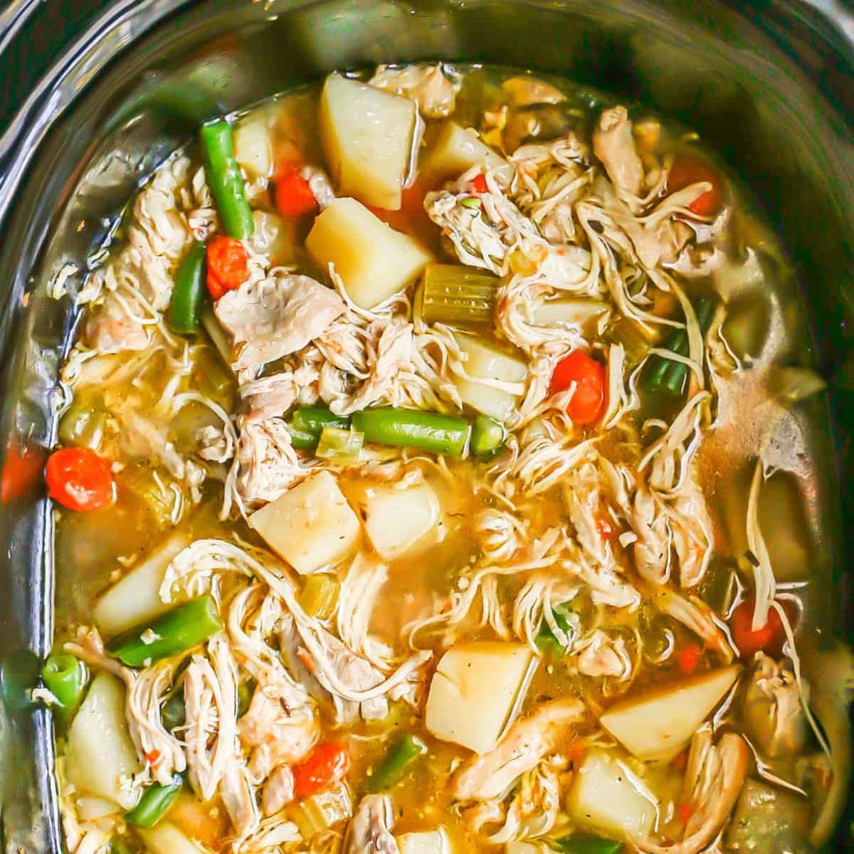 A slow cooker insert filled with chicken stew with potatoes, green beans, carrots and shredded chicken.