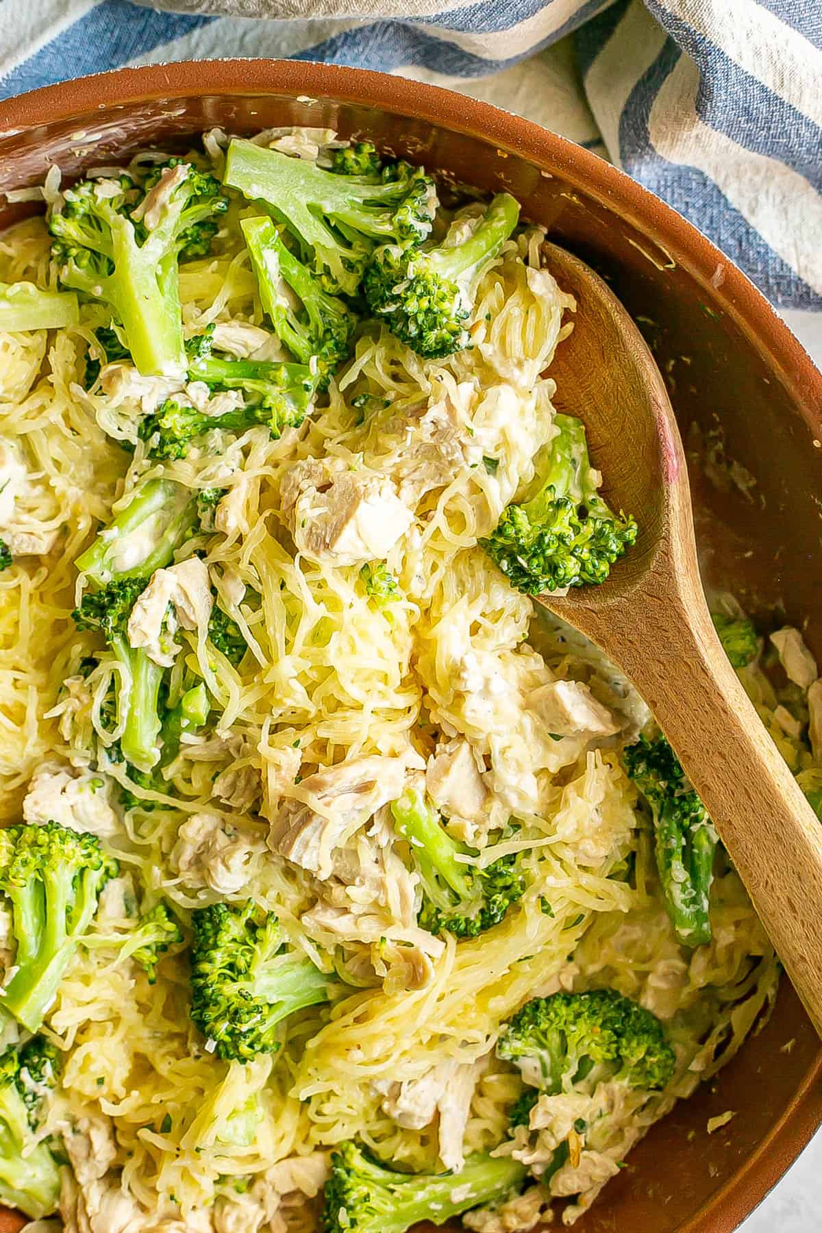 A wooden spoon resting in a skillet of spaghetti squash, chicken and broccoli with a light creamy sauce.