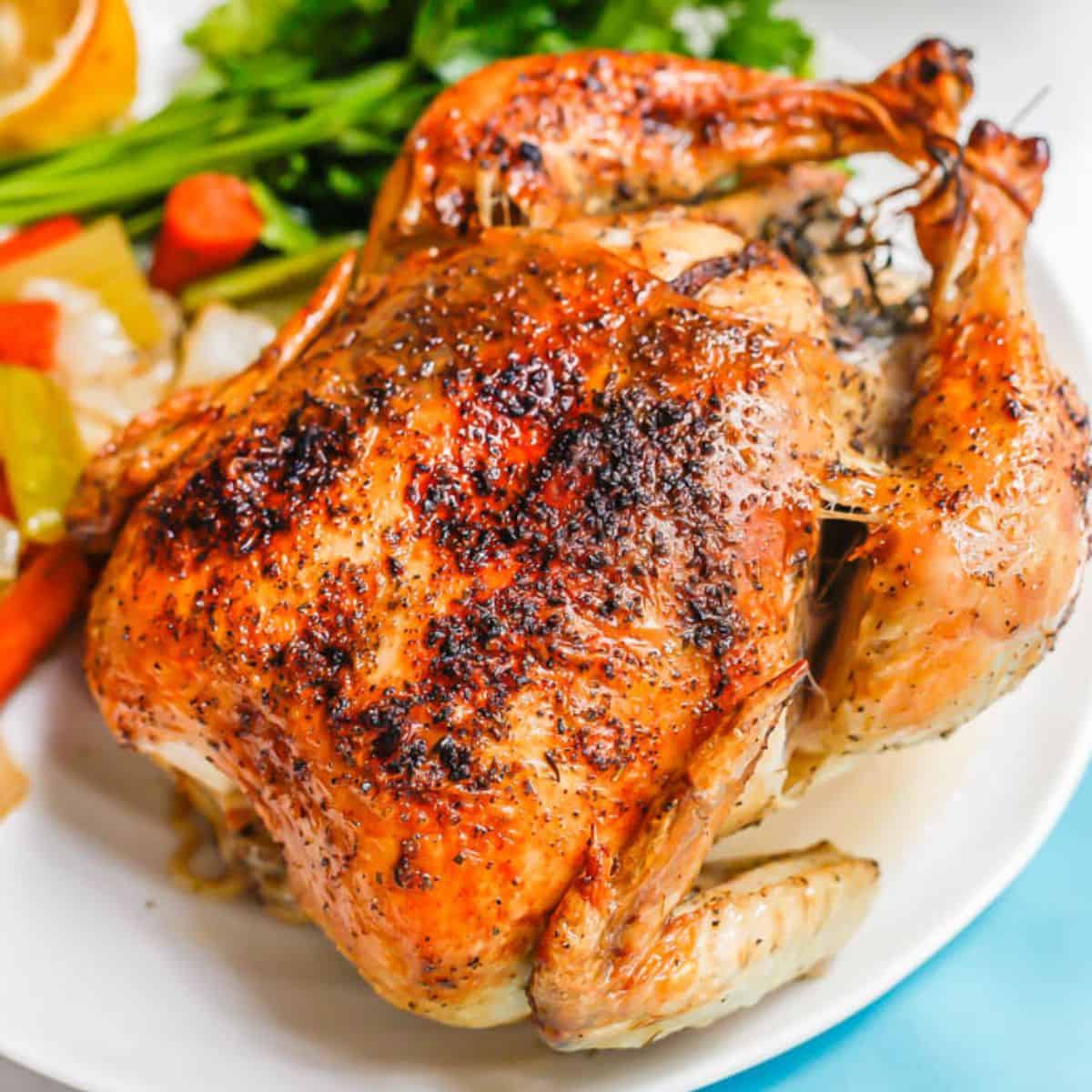 Close up of a whole roasted chicken on a large white serving platter with veggies and fresh parsley to the side.