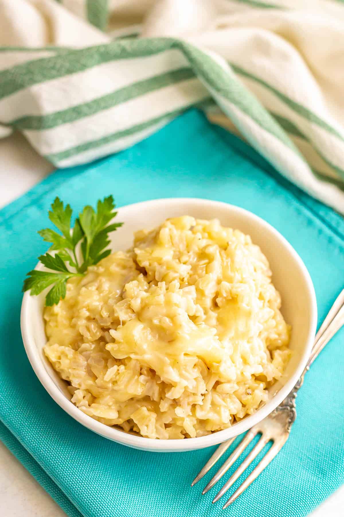 A bowl of cheesy brown rice with a garnish of parsley on the side and a fork nearby on a teal napkin.