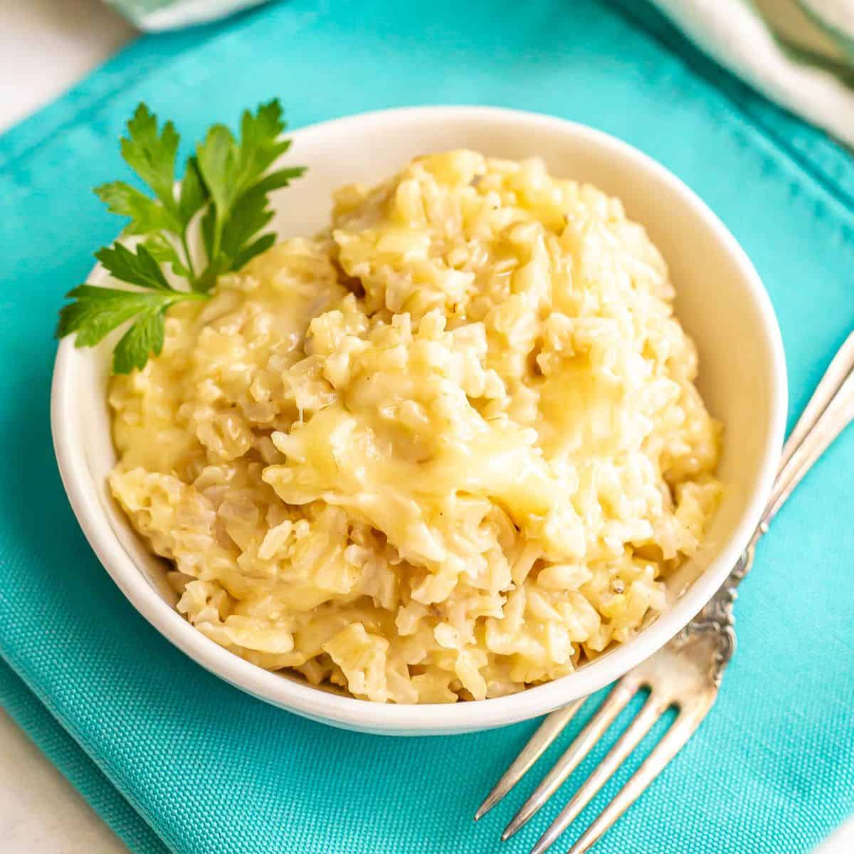 Close up of a bowl of cheesy brown rice with a garnish of parsley on the side and a fork nearby on a teal napkin.