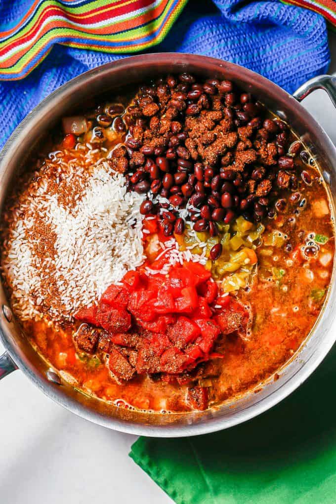 A large deep skillet with black beans, tomatoes, diced green chilies and white rice, plus seasonings, arranged before being stirred together.