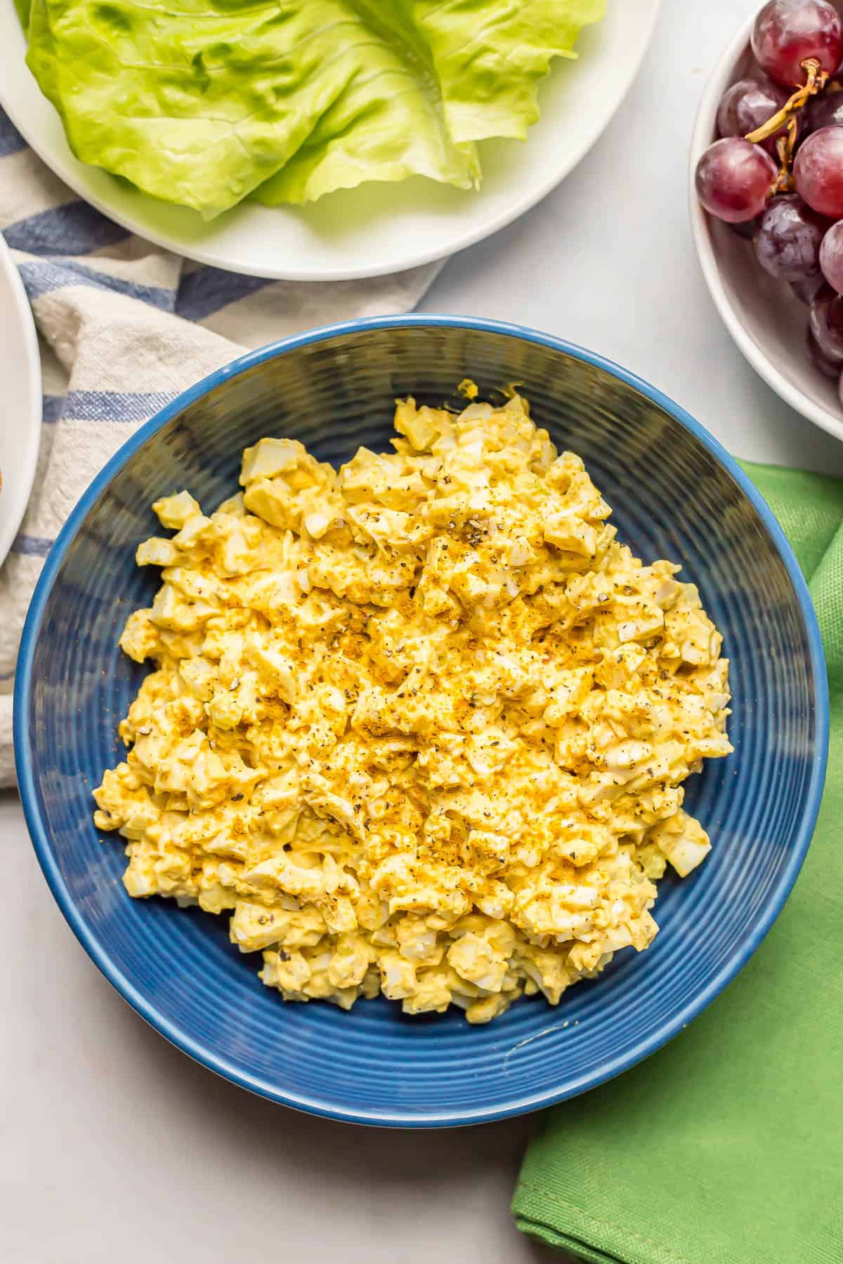 A blue bowl filled with curried egg salad with plates of lettuce and red grapes to the side.