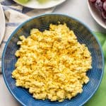 A blue bowl filled with curried egg salad with plates of lettuce and red grapes to the side and a text overlay on the photo.