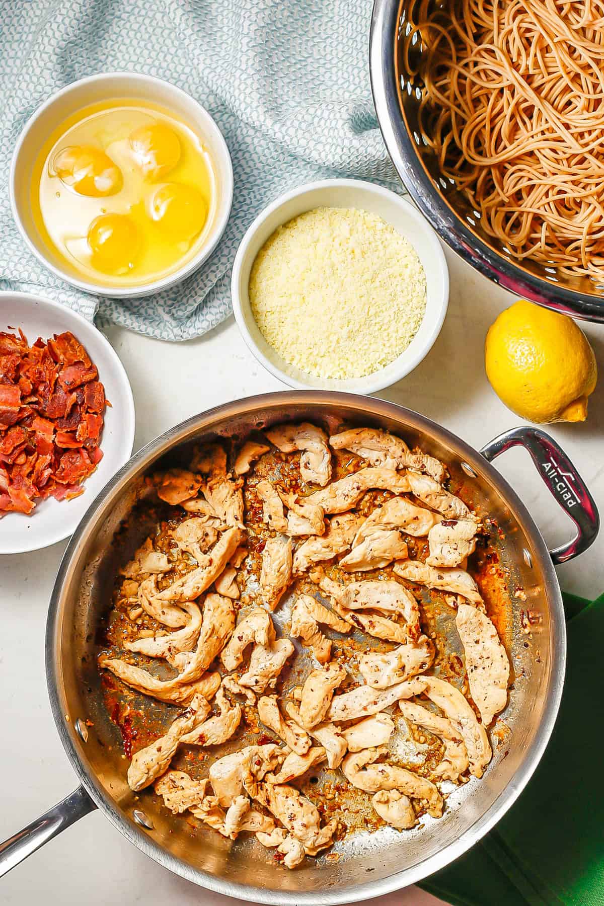 Seared chicken in a pan, drained cooked spaghetti noodles in a colander and bowls of bacon, eggs and Parmesan cheese laid out on a counter.