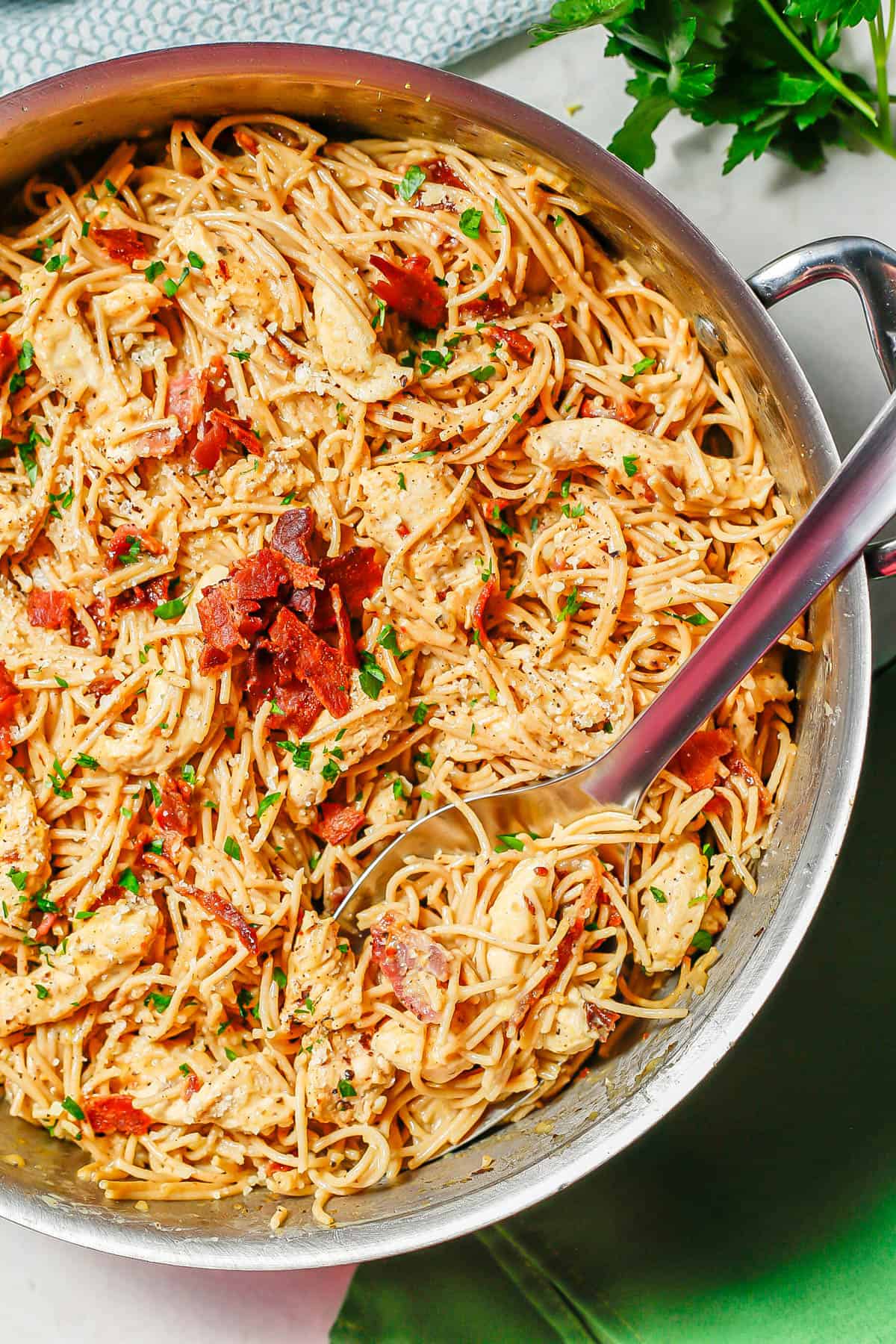 A large silver spoon resting in a deep skillet with chicken carbonara topped with bacon and parsley.